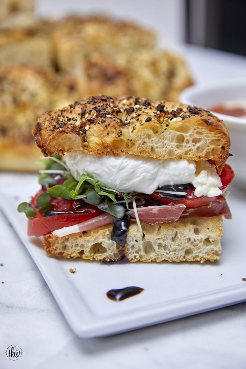 Herbaceous crunchy no-knead focaccia topped with prosciutto, tomatoes, balsamic glaze, microgreens, creamy burrata, olive oil & sea salt flakes making it one of the best sandwiches you'll ever sink your teeth into! Indulge in a Scrumptious Prosciutto, Tomato, & Burrata Focaccia Sandwich, no knead focaccia, Italian sandwich, burrata sandwich, crusty bread